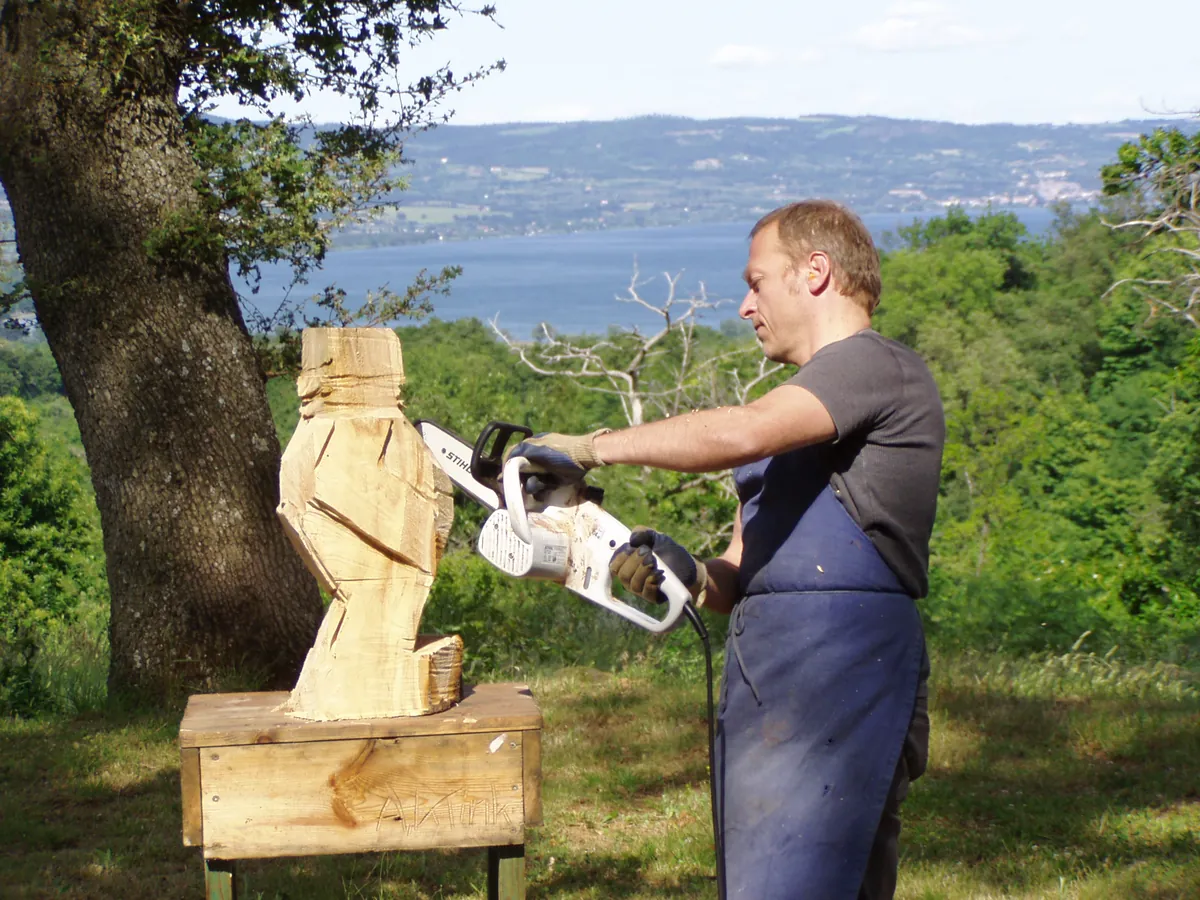Albrecht Klink arbeitet mit der Motorsäge an einer Holzskulptur, im Hintergrund der Bolsenasee im nördlichen Latium