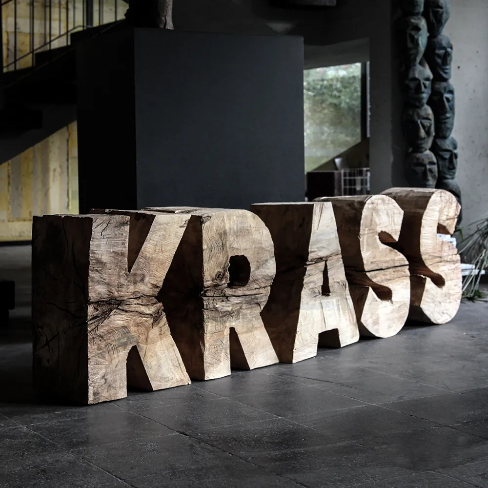 Die Buchstaben KRASS, aus einem Pappelstamm geschnitzt, als Holzskulptur in einem Raum aus Beton